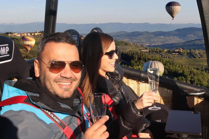 Couple in hot air balloon drinking champagne, scenic view with other balloons in background.