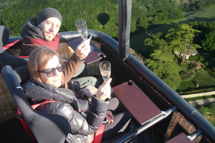 Two people in a hot air balloon, toasting with champagne glasses over a green landscape.